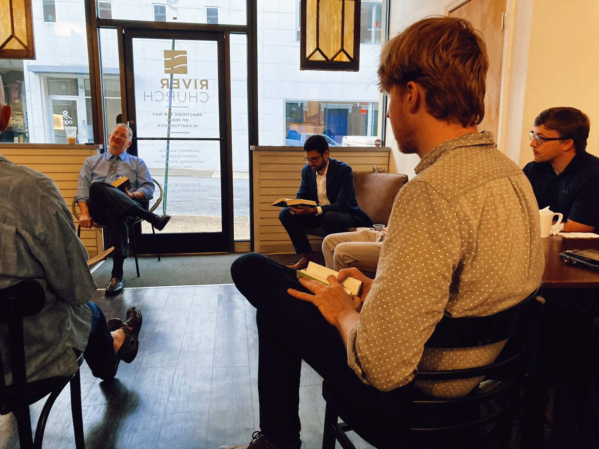 Men seated in armchairs reading books in a quiet room with a glass front door